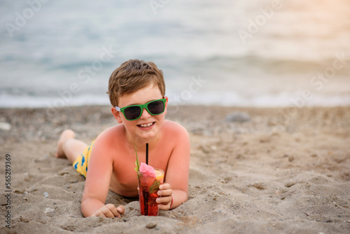 фотографія Little boy kid with sunglasses is lying on the sand near seaside smiling and drinking a cocktail