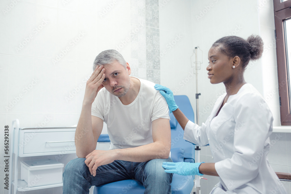 Fototapeta premium An unhappy adult man suffering from pain, severe headache, stress, ocular migraine, sits in a room and asks a doctor for a check and help in treatment.