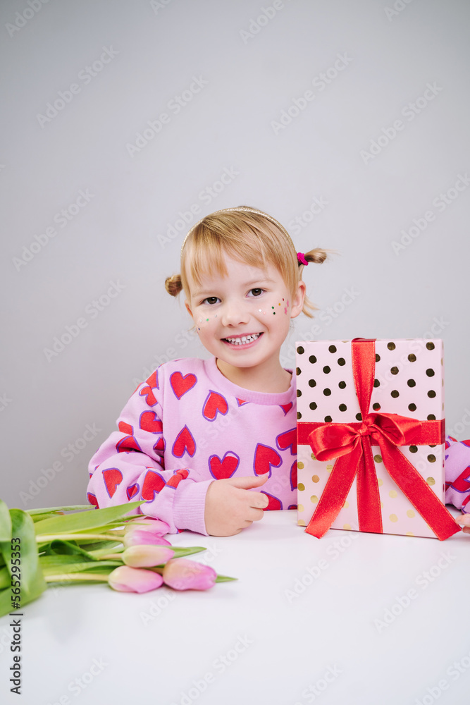 Cheerful little girl in pink sweatshirt with red hearts pattern with bouquet of pink tulips and gift box isolated over gray white background.