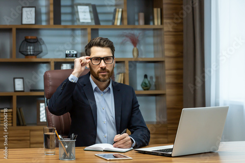 Wallpaper Mural Serious thinking boss inside the office portrait of a mature businessman with a beard and glasses, the man is looking at the camera with concentration, working with documents on a laptop at the table Torontodigital.ca