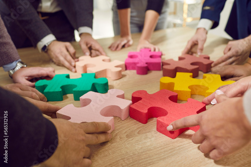 Business team connecting different jigsaw puzzle pieces. Group of young office workers joining colorful parts on wooden table. Cropped shot of human hands holding pieces. Teamwork concept background