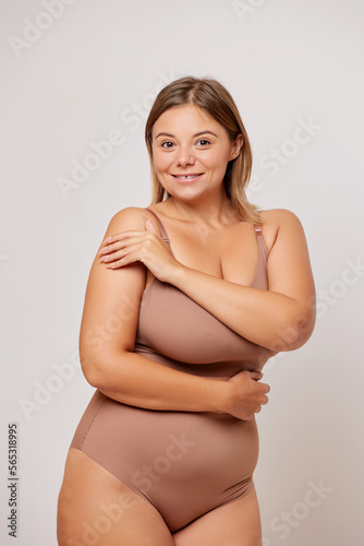 Self acceptance concept. Vertical shot of body positive a sensual woman in her 30s feels comfortable in her body and self loved, hugs herself, poses in beige lingerie against white background © South House Studio