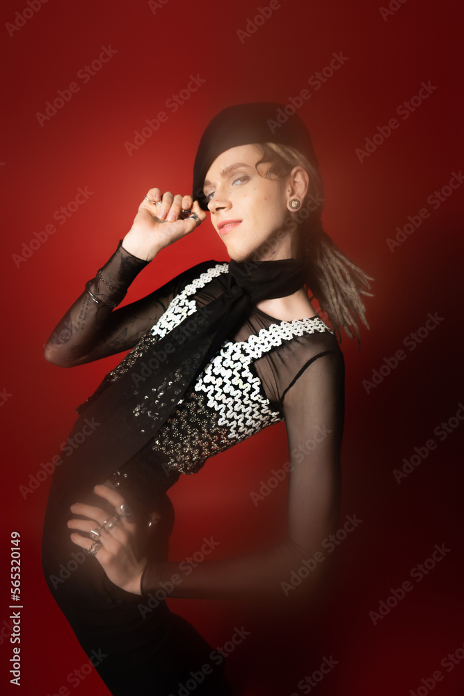 queer person in elegant attire adjusting black beret while posing with hand on hip on red background. queer person in elegant attire adjusting black beret while posing with hand on hip on red background.