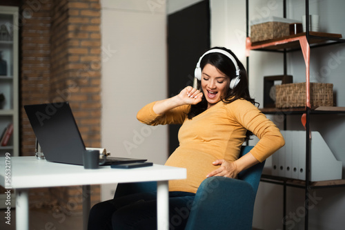 Beautiful pregnant listening the music while working on laptop. Young businesswoman working in her office.