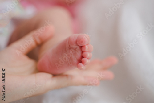 Close up mother holding foot newborn girl in a room. Adorable infant rests on white bedsheets, staring at camera looking peaceful. Infancy, healthcare and paediatrics, babyhood concept.