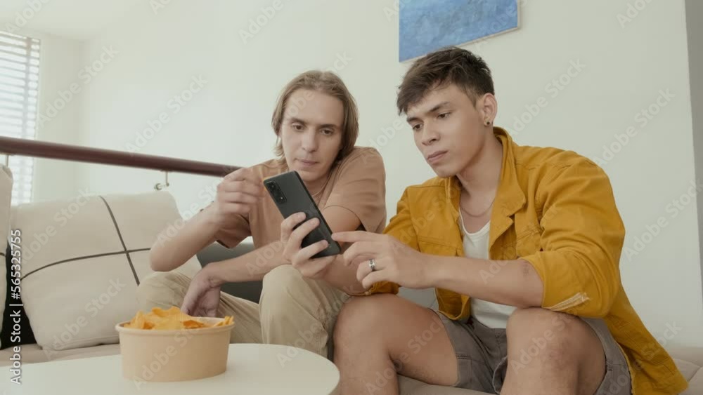 Two buddies eating chips and discussing something on smartphone sitting on couch at home Two buddies eating chips and discussing something on smartphone sitting on couch at home