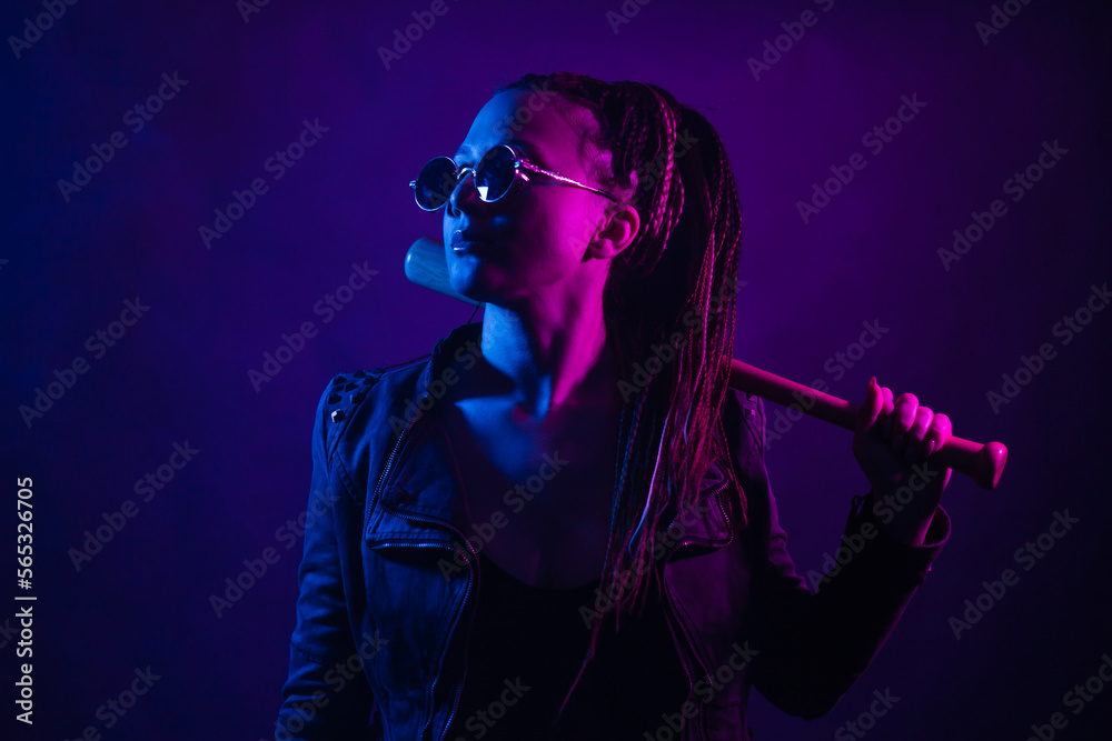 A young woman with dreadlocks in the dark in a club in black glasses holds a painless bat in her hands. Girl with afro-braids at night in neon color.