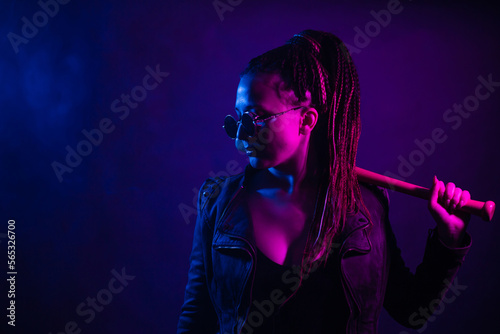 A young woman with dreadlocks in the dark in a club in black glasses holds a painless bat in her hands. Girl with afro-braids at night in neon color.