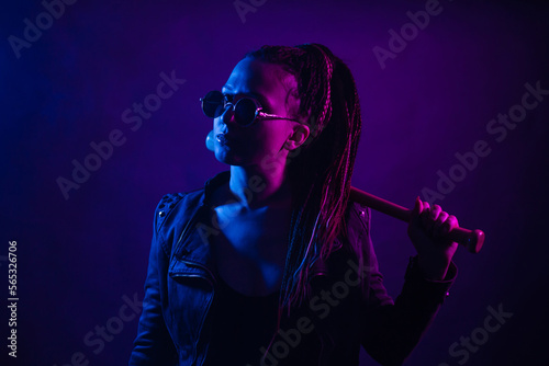 A young woman with dreadlocks in the dark in a club in black glasses holds a painless bat in her hands. Girl with afro-braids at night in neon color.