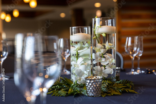 Fotografía A centerpiece at a wedding with wine glasses on the table, flowers and floating candles in pillar vases, and blurred out lights in the distance