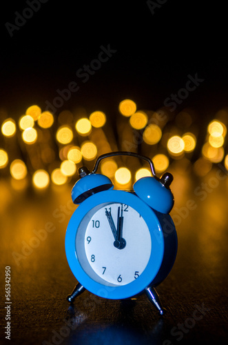 Classic arrow blue alarm clock on black background, illuminated from the side, behind round yellow blurred lights