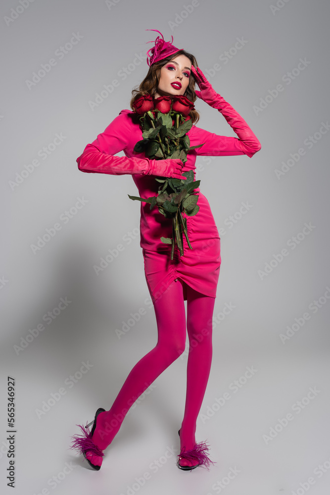 full length of young woman in magenta color gloves and hat with feather holding red roses on grey. full length of young woman in magenta color gloves and hat with feather holding red roses on grey.