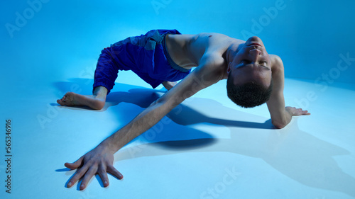 Contemporary dance style. Young shirtless man dancing over blue studio background in neon. Contemp, experimental. Concept of art, body aesthetics, motion, action, inspiration.