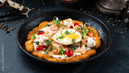 Fried big potato pancake with salmon fish, tomatoes, egg, onion, herbs and sour cream in a frying pan.