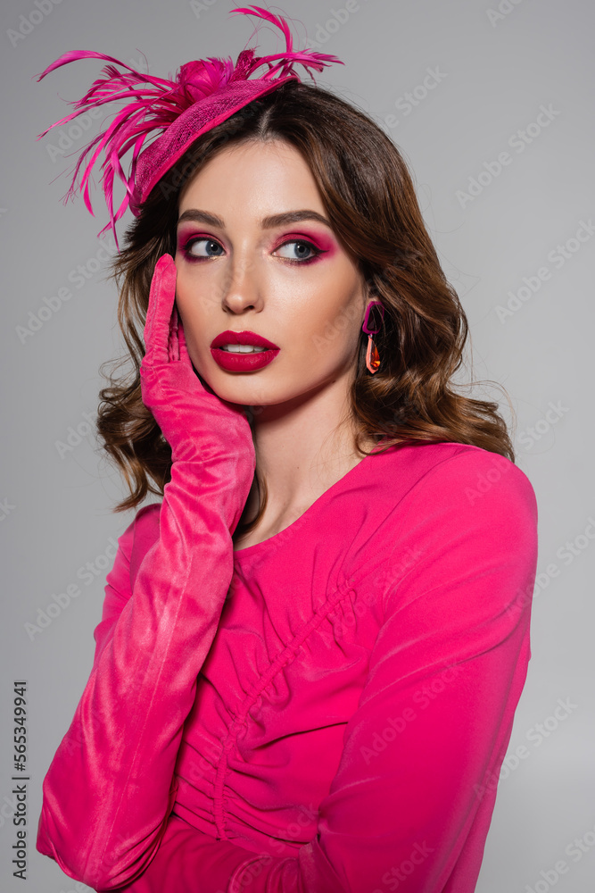 dreamy young woman in magenta color dress and elegant hat with feather looking away isolated on grey.