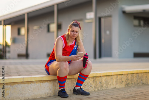 Female football player holding water and checking phone, sitting down after game