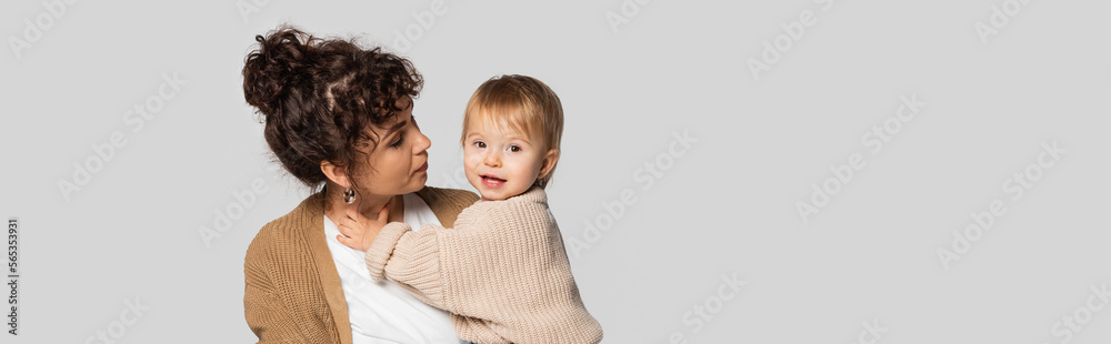 stylish mother in casual clothes holding in arms cheerful baby girl in sweater isolated on grey, banner.