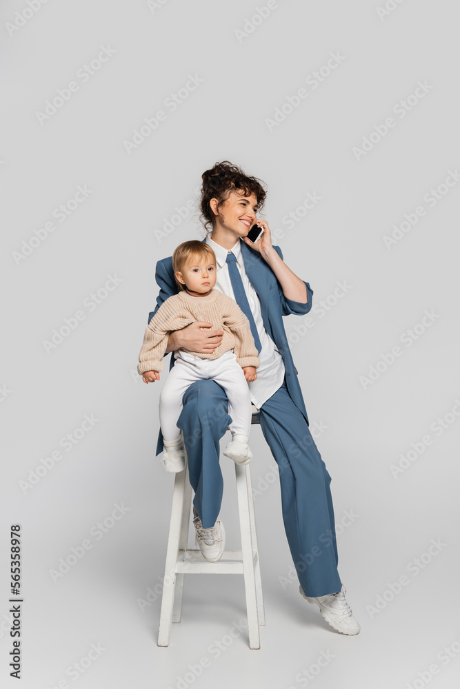 happy businesswoman in blue suit talking on smartphone and sitting with toddler daughter on high chair on grey. happy businesswoman in blue suit talking on smartphone and sitting with toddler daughter on high chair on grey.