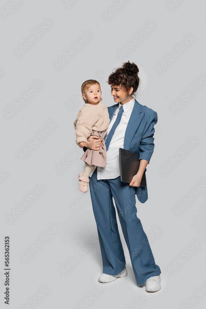 full length of cheerful businesswoman in blue suit holding in arms toddler daughter and laptop on grey. full length of cheerful businesswoman in blue suit holding in arms toddler daughter and laptop on grey.
