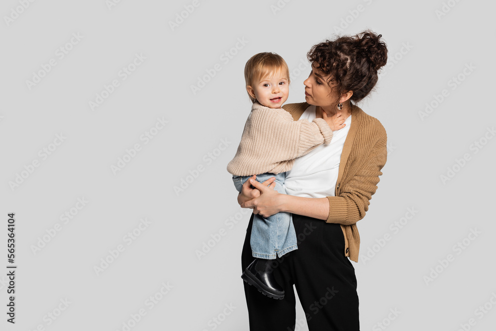 stylish mother in casual clothes holding in arms cheerful baby girl in sweater isolated on grey. stylish mother in casual clothes holding in arms cheerful baby girl in sweater isolated on grey.