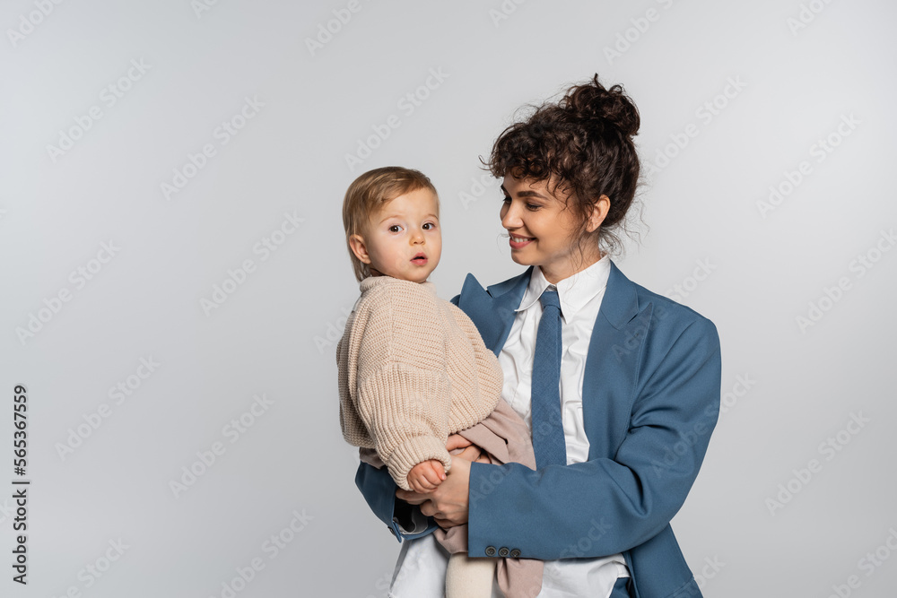 happy businesswoman in suit with tie looking at baby girl isolated on grey. happy businesswoman in suit with tie looking at baby girl isolated on grey.