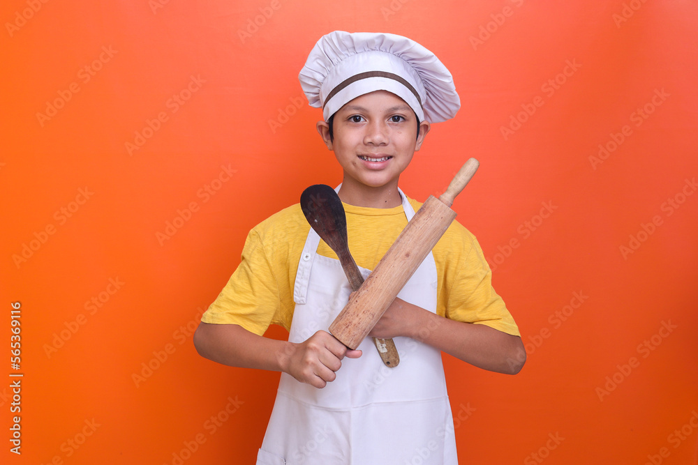 Smiling little boy in chef's toque and apron holding wooden rolling pin and spatula on orange background. Smiling little boy in chef's toque and apron holding wooden rolling pin and spatula on orange background.