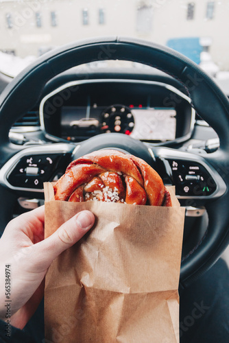 Delicious cinnamon bun in hand in front of a steering wheel of a car