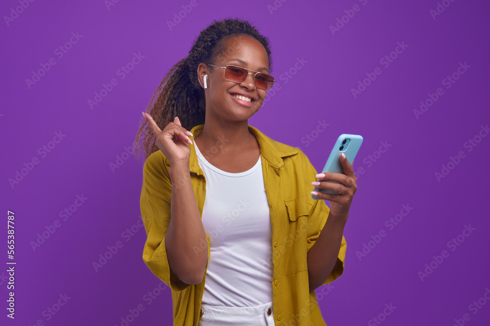 Young confident happy African American woman smiles broadly holding phone in hands and pointing finger up enjoys using cool headphones stands on isolated purple background. Music, meloman, playlist Young confident happy African American woman smiles broadly holding phone in hands and pointing finger up enjoys using cool headphones stands on isolated purple background. Music, meloman, playlist
