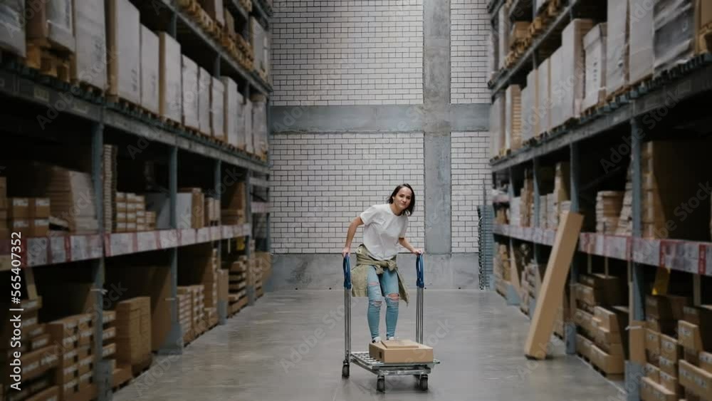 Girl rides shopping cart in furniture store warehouse between the rows of shelves with cardboard boxes Girl rides shopping cart in furniture store warehouse between the rows of shelves with cardboard boxes