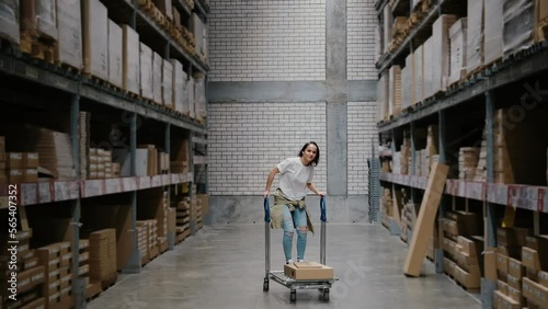 Girl rides shopping cart in furniture store warehouse between the rows of shelves with cardboard boxes