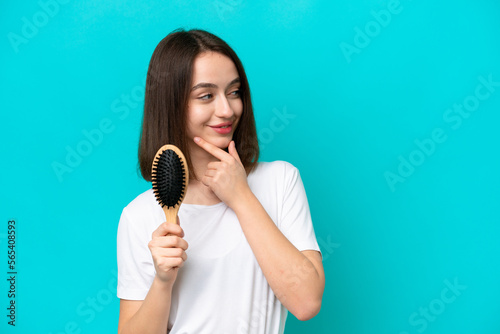 Young ukrainian woman with hair comb isolated on blue background thinking an idea and looking side