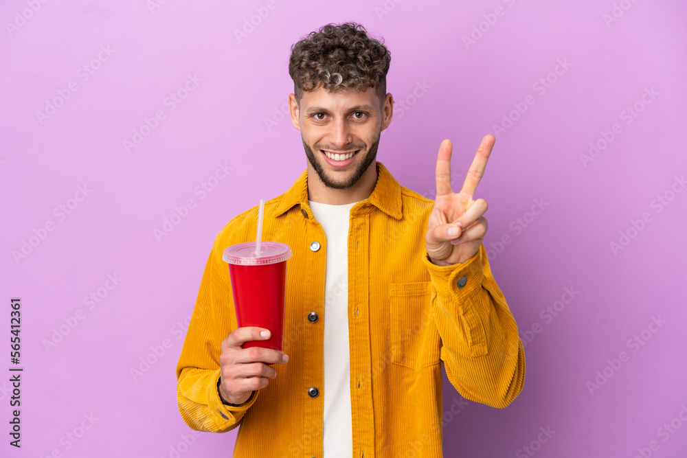Young blonde man holding soda isolated on purple background smiling and showing victory sign Young blonde man holding soda isolated on purple background smiling and showing victory sign