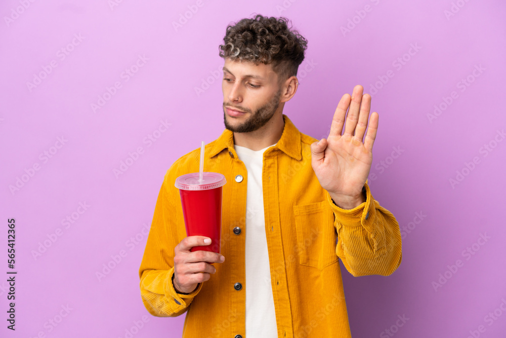 Young blonde man holding soda isolated on purple background making stop gesture and disappointed Young blonde man holding soda isolated on purple background making stop gesture and disappointed