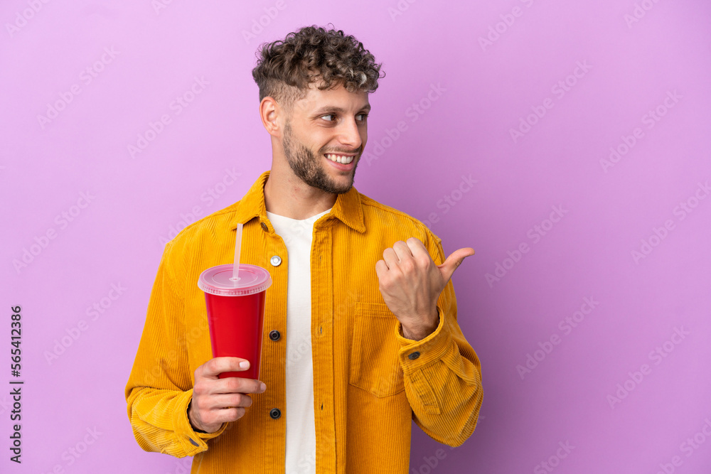 Fototapeta premium Young blonde man holding soda isolated on purple background pointing to the side to present a product
