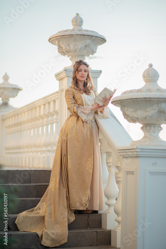 A beautiful young woman in an eighteenth century historical gold dress stands on the stairs of a mansion with a book in her hands. the princess is reading in the palace.