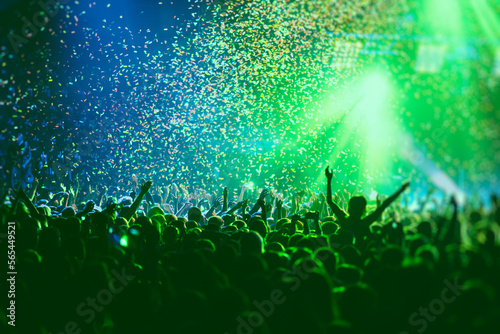 A crowded concert hall with scene stage green lights, rock show performance, with people silhouette, colourful confetti explosion fired on dance floor air during a concert festival
