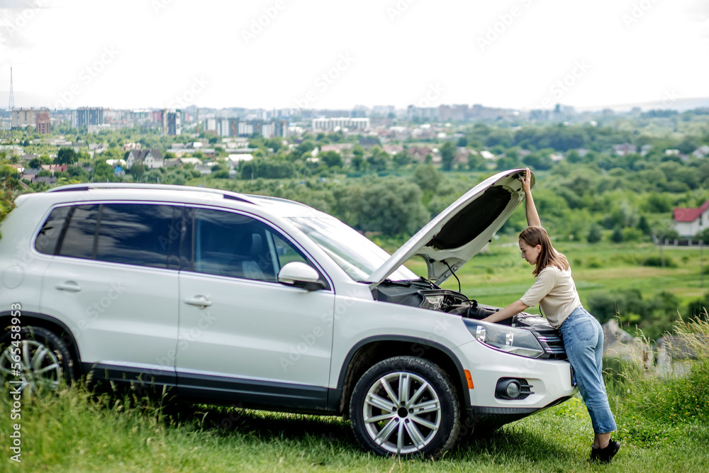 Confused female driver standing outside her car with the hood up and looking for the cause of the car breakdown