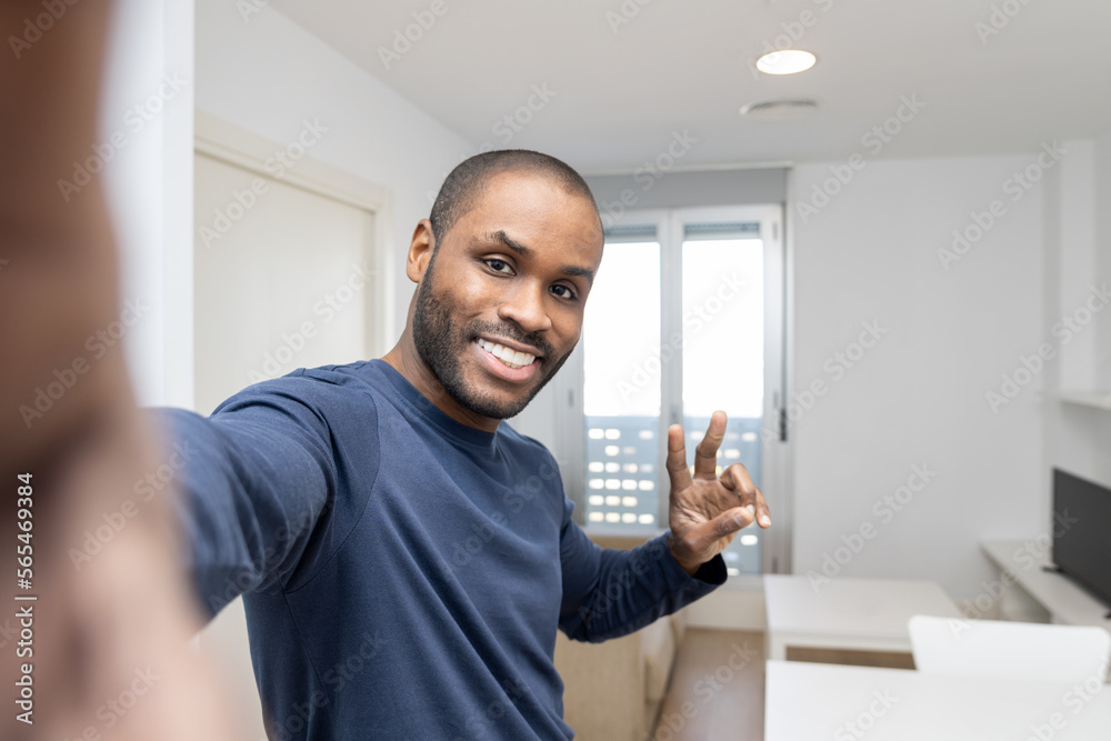 A dark-skinned man takes a selfie in front of a modern smartphone camera with the gesture of victory against the background of a new white apartment he rented, celebrating the last mortgage payment. A dark-skinned man takes a selfie in front of a modern smartphone camera with the gesture of victory against the background of a new white apartment he rented, celebrating the last mortgage payment.