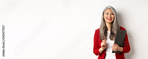 Successful asian lady boss in red blazer, holding clipboard with documens and pen, working and looking happy, white background photo