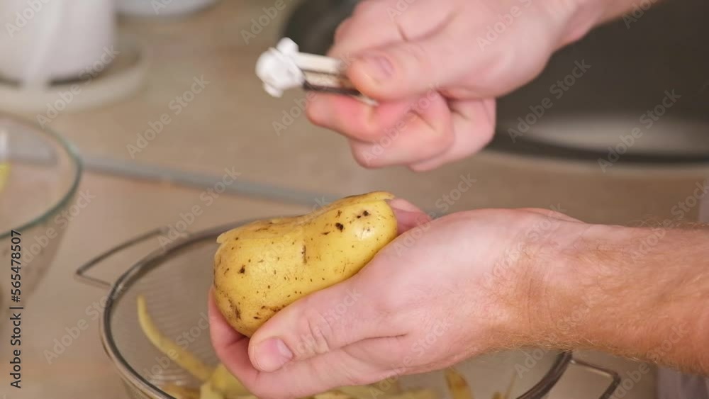 A man peels potatoes with a vegetable peeler in a bright kitchen. Preparation of potatoes for cooking. Cleaning the peel from pesticides. A man cooks dinner or lunch at home for family. A man peels potatoes with a vegetable peeler in a bright kitchen. Preparation of potatoes for cooking. Cleaning the peel from pesticides. A man cooks dinner or lunch at home for family.