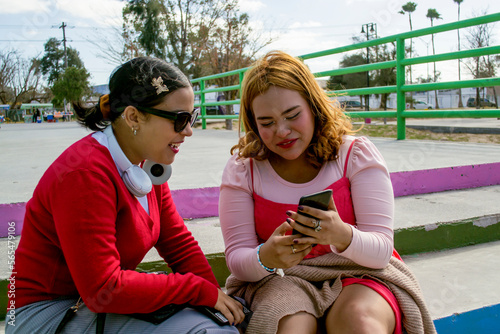 Chicas jóvenes viendo videos en teléfono celular sentadas en un parque al aire libre. Mujeres latinas