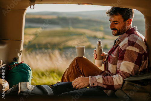 Man resting after a long walk in a car trunk, sipping a hot tea, and enjoying a sunset view.