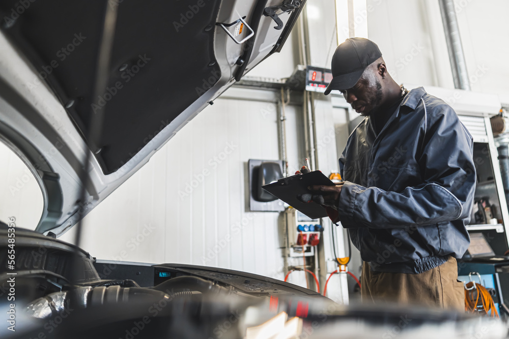 Skilled car mechanic inspects the car engine at the repair facility and takes notes. High-quality photo