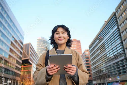 Portrait of happy korean girl smiles, walks in city with digital tablet and backpack, uses her gadget to get around photo