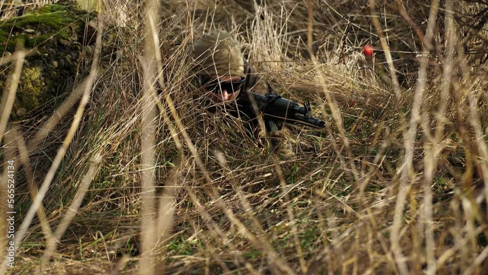 The soldier lies on the ground and disguises himself in the dry grass. A military man holds a firearm in his hands and is dressed in an armored uniform. Military operation. The concept of war. The soldier lies on the ground and disguises himself in the dry grass. A military man holds a firearm in his hands and is dressed in an armored uniform. Military operation. The concept of war.