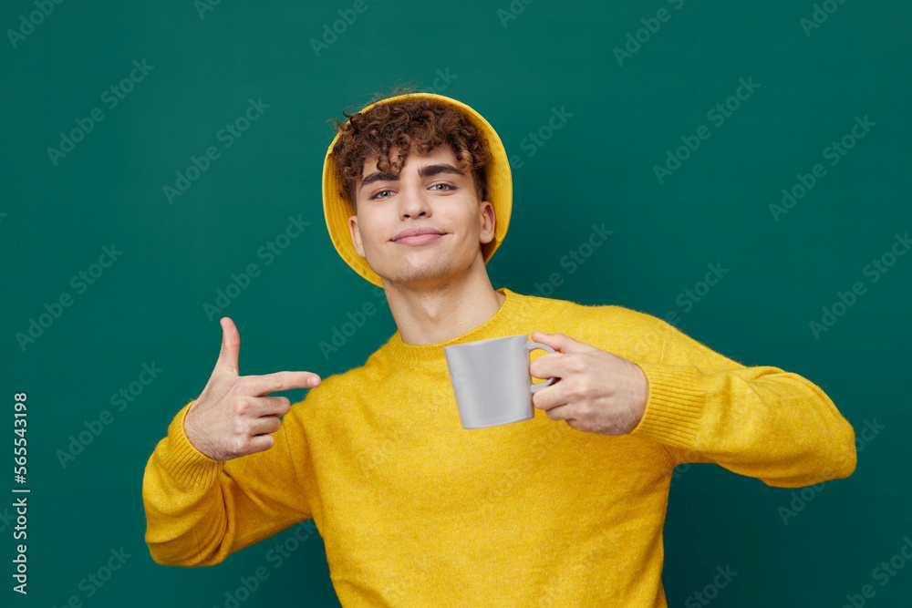 a handsome man stands on a green background in a yellow T - shirt and hat holding a metal glass in his hand pointing at it with his finger . Horizontal photo for inserting an advertising layout