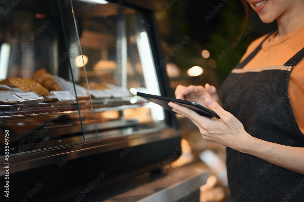 Smiling coffee shop owner using digital tablet to receiving online orders while standing near showcase with cakes and desserts Smiling coffee shop owner using digital tablet to receiving online orders while standing near showcase with cakes and desserts