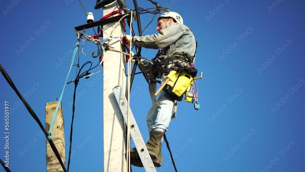 Skilled electrician in helmet fixes wires standing on ladder near high pole against blue sky on summer day backside view. Electrical service and mounting on the pole. Slow motion Skilled electrician in helmet fixes wires standing on ladder near high pole against blue sky on summer day backside view. Electrical service and mounting on the pole. Slow motion