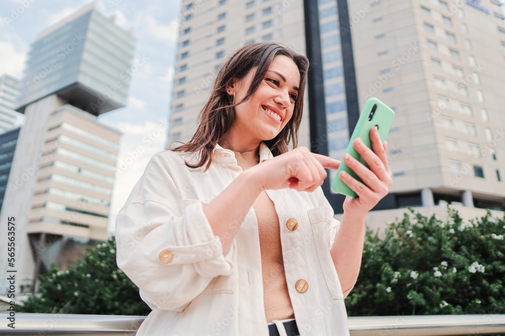 Naklejka premium Happy young woman touching the screen of the cell phone with her finger, scrolling and swipe up the internet app. Brunette teenager smiling using the smartphone outdoors in front of city buildings