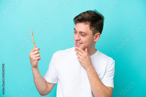 Young Brazilian man brushing teeth isolated on blue background looking to the side and smiling